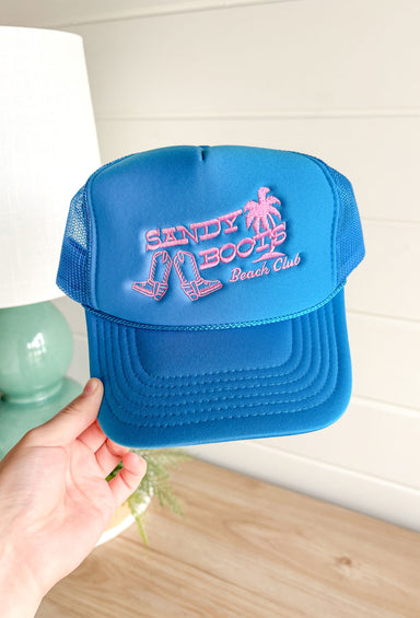 Charlie Southern: Sandy Boots Trucker Hat, Cobalt blue otto trucker hat with embroidered boots and a palm tree with text "sandy boots beach club" in pink