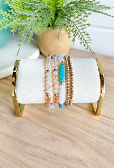 Seeking Sunshine Bracelet Set, stack of 5 bracelets, two of them are clear, tan, and gold beads; one of them is milky white, clear, teal, and gold beads; and two are all gold beaded bracelets 