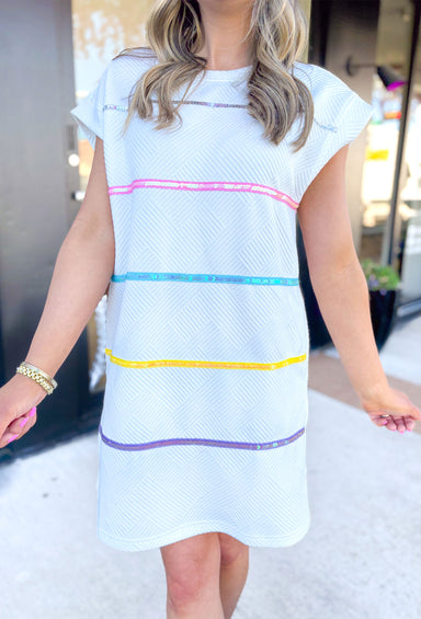 All The Attention Dress in White, short sleeve white textured dress with pockets, silver, baby pink, electric light blue, yellow, & purple horizontal sequin stripes on the dress