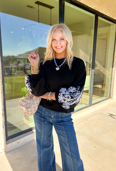 Standout sleeves make this black pullover a fall favorite. Designed with bold white embroidery, the pattern brings striking contrast and texture, while the ribbed cuffs create a softly billowed effect at the wrists. A ribbed hem completes the look, giving the sweater a polished yet cozy finish. Made from a soft cotton-blend knit, it’s perfect for layering into your everyday wardrobe while keeping that touch of bold, artistic flair.