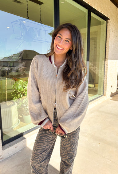 Cozy and casual, this tan knit zip-up sweater is the perfect throw-on-and-go piece for fall. Designed with burgundy and off-white trim around the neck and sleeves, it adds just the right amount of contrast to keep the look fresh. Front pockets bring a practical touch, while the soft knit blend makes it warm yet lightweight for layering. Whether styled with jeans for everyday wear or joggers for lounging, this sweater is a must-have seasonal staple.