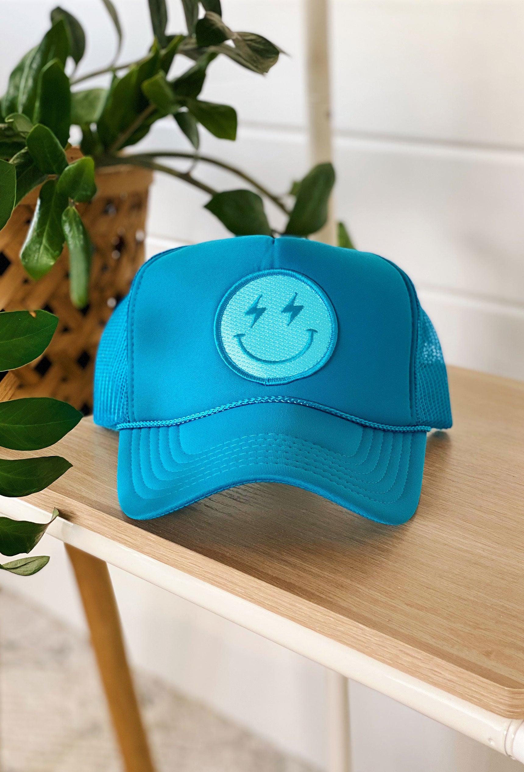 Happy Vibes Trucker Hat, turquoise blue, mesh backing, adjustable strap, blue smiley face patch with lightening bolts as eyes